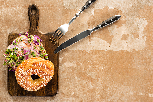 top view of fresh delicious bagel with meat, red onion and sprouts on wooden cutting board with cutlery on aged beige surface