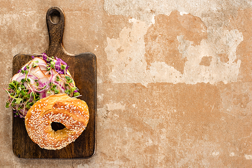 top view of fresh delicious bagel with meat, red onion and sprouts on wooden cutting board on aged beige surface