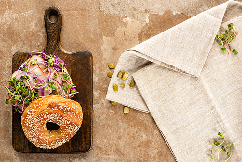 top view of fresh delicious bagel with meat, red onion and sprouts on wooden cutting board near napkin on aged beige surface
