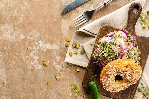 top view of fresh delicious bagel with meat, red onion and sprouts on wooden cutting board near napkin and cutlery on aged beige surface