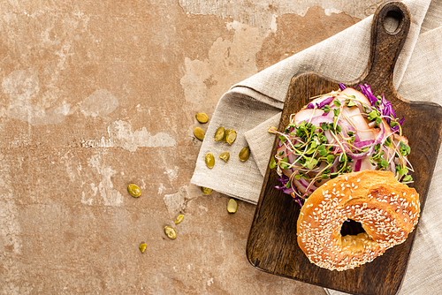 top view of fresh delicious bagel with meat, red onion and sprouts on wooden cutting board on napkin with pumpkin seeds on aged beige surface