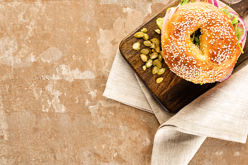 top view of fresh delicious bagel on wooden cutting board with pumpkin seeds on aged beige surface with napkin