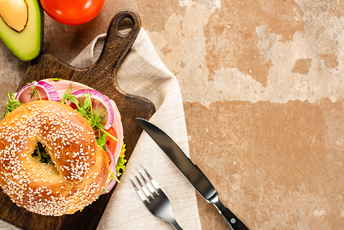 top view of fresh delicious bagel on wooden cutting board on aged beige surface with s, cutlery and napkin