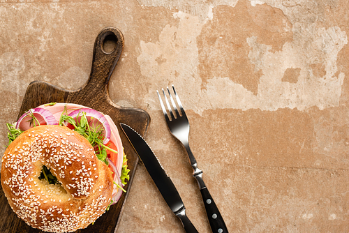 top view of fresh delicious bagel on wooden cutting board on aged beige surface with cutlery