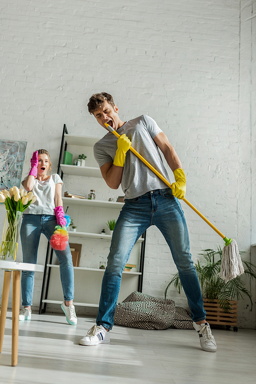 young and surprised woman looking at handsome man singing while holding mop