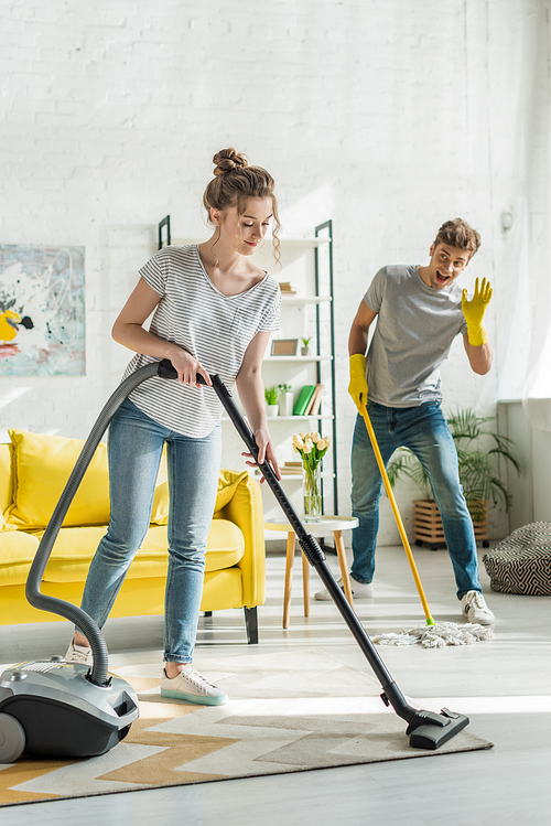 attractive girl using vacuum cleaner near man waving hand while washing floor with mop