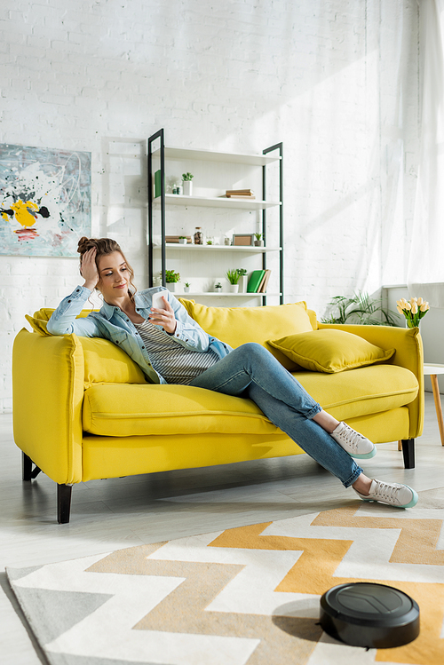 attractive girl using smartphone while robotic vacuum cleaner washing carpet in living room