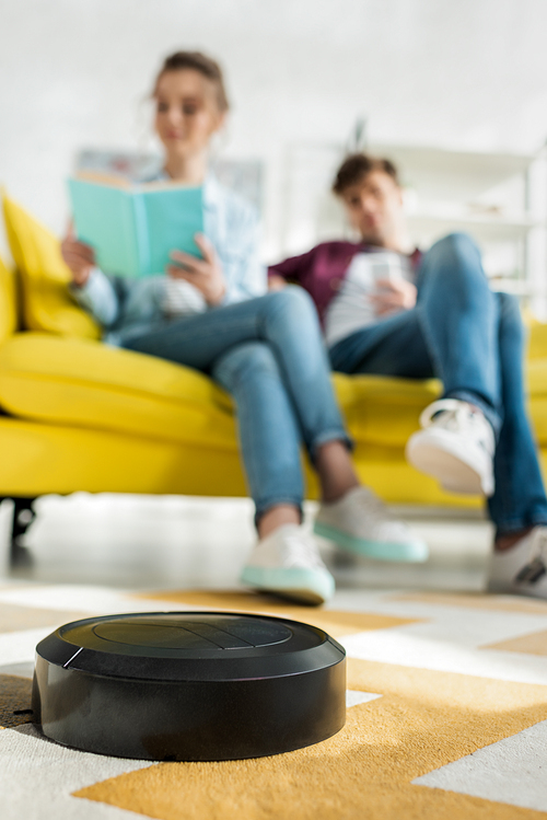 selective focus of robotic vacuum cleaner washing carpet near man and woman in living room