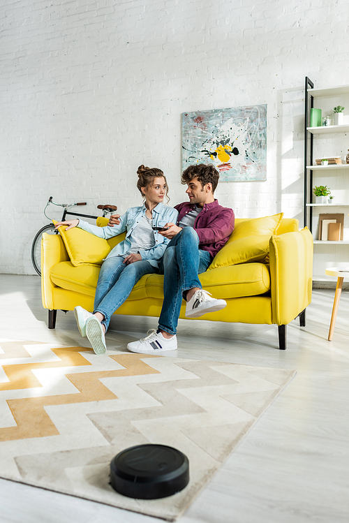 happy man and woman watching movie while robotic vacuum cleaner washing carpet in living room