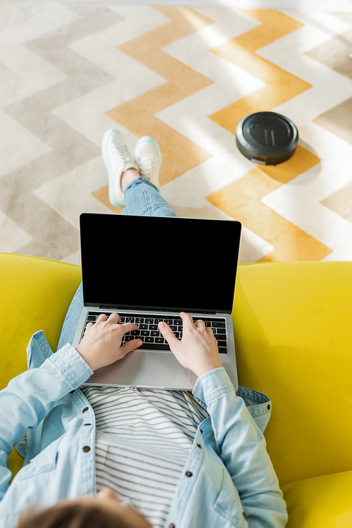 top view of girl using laptop with blank screen while robotic vacuum cleaner washing carpet in living room