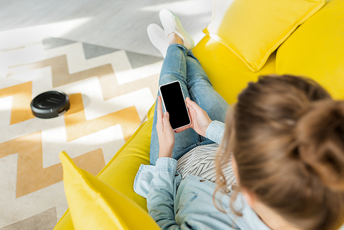 top view of woman holding smartphone with blank screen while robotic vacuum cleaner washing carpet in living room