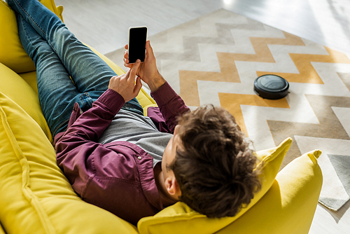 overhead view of man relaxing on sofa and holding smartphone with blank screen while robotic vacuum cleaner washing carpet in living room