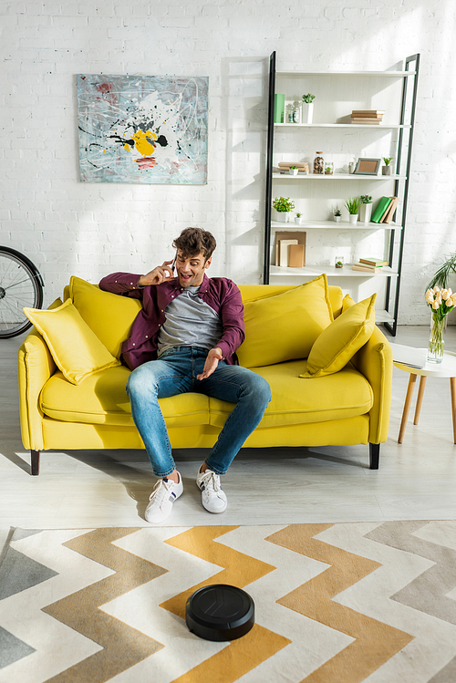 happy man talking on smartphone while robotic vacuum cleaner washing carpet in living room