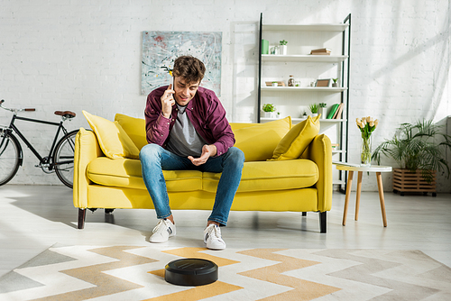 cheerful man talking on smartphone while robotic vacuum cleaner washing carpet in living room