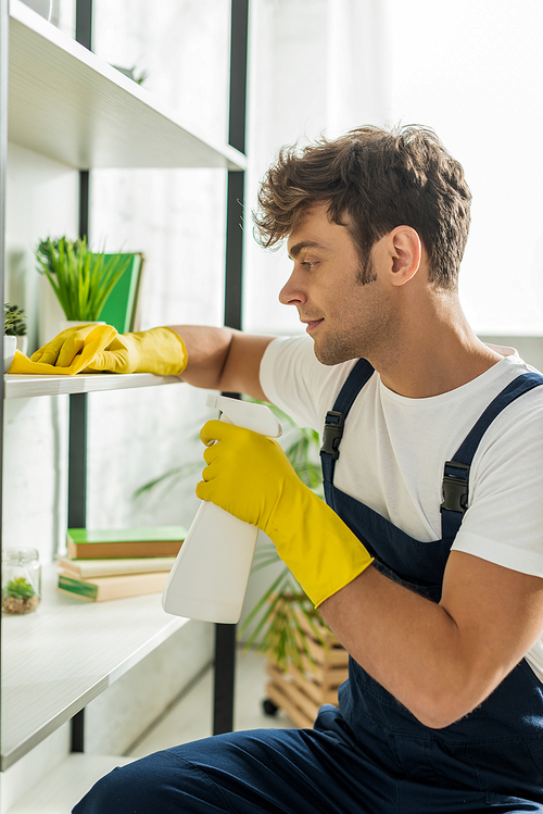 side view of handsome man in overalls cleaning rack shelves in apartment