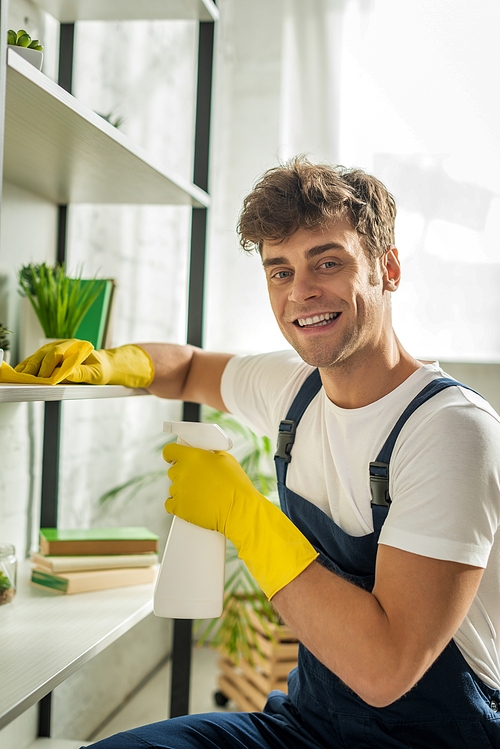 cheerful cleaner in overalls cleaning rack shelves in apartment