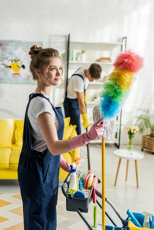 selective focus of attractive cleaner holding duster brush near cleaning trolley