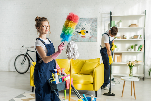 selective focus of young and happy cleaner holding duster brush near cleaning trolley
