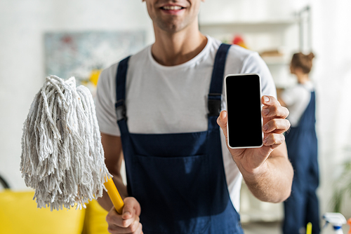 cropped view of happy cleaner holding smartphone with blank screen and mop