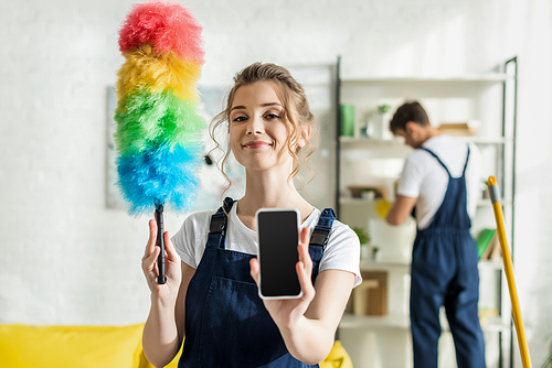selective focus of happy and attractive cleaner holding smartphone with blank screen and duster brush