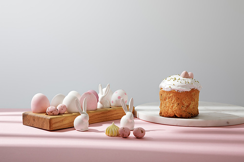 Easter eggs on wooden board with decorative rabbits near easter bread on pink and grey background