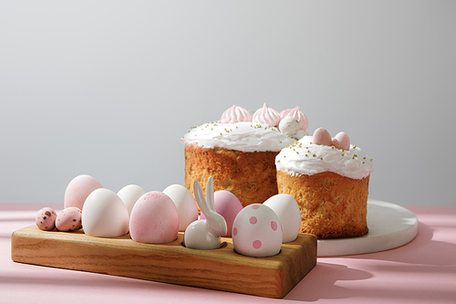 Easter eggs on wooden board with decorative rabbit near easter cakes on pink and grey background