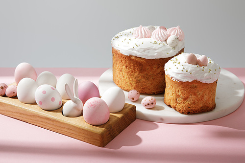 Easter eggs on wooden board with decorative bunny near easter cakes on pink and grey