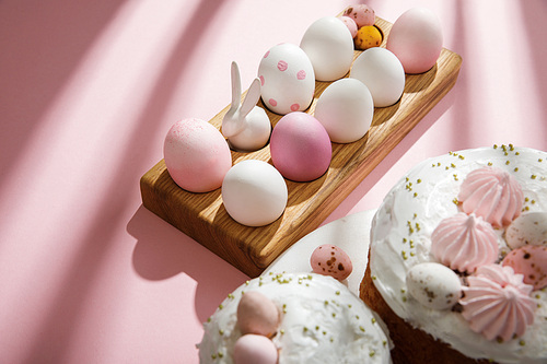 Selective focus of painted chicken and quail eggs, decorative bunny on wooden board near easter cakes on pink background