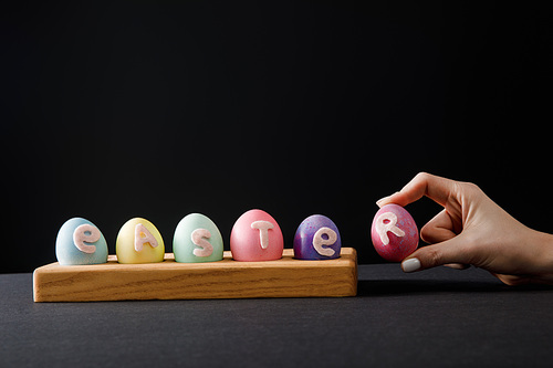 Cropped view of woman holding painted chicken egg near board with colorful eggs with easter lettering on grey and black
