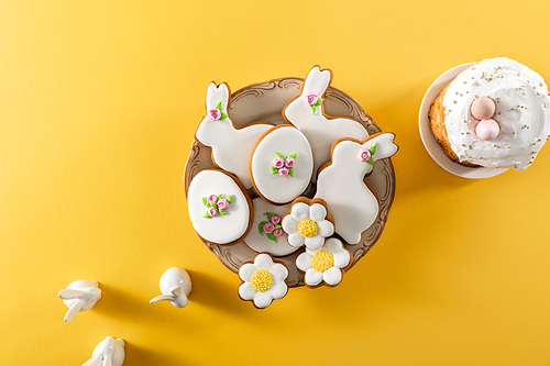 Top view of bowl with tasty cookies, decorative bunnies and easter cake on yellow background