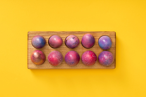 Top view of wooden board with colorful Easter eggs on yellow background