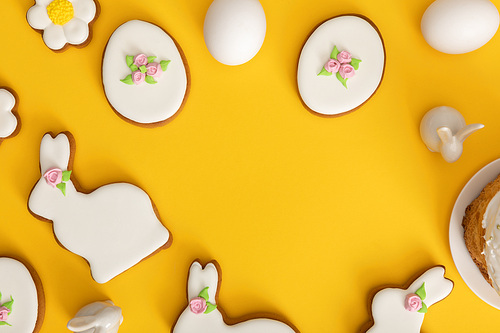 Top view of chicken eggs, cookies and decorative bunnies on yellow background