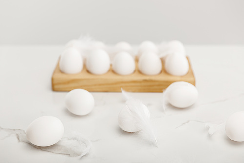 Selective focus of chicken eggs on wooden egg tray and feathers on grey