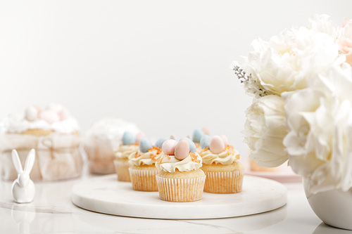 Selective focus of cupcakes on round board, decorative bunny and vase with flowers on white background