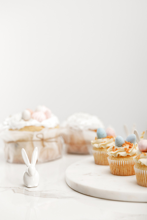 Selective focus of cupcakes on round board, decorative bunny and easter cakes on grey background