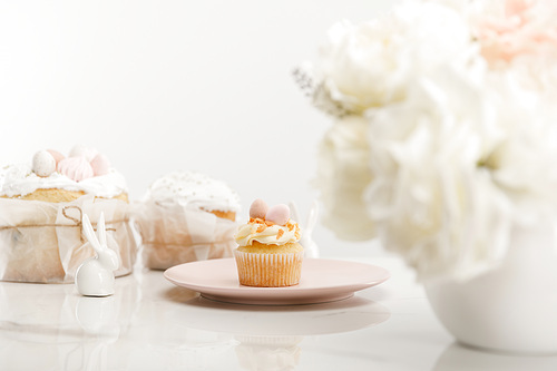selective focus of cupcake on plate, decorative rabbits, . cakes and vase with bouquet on white background