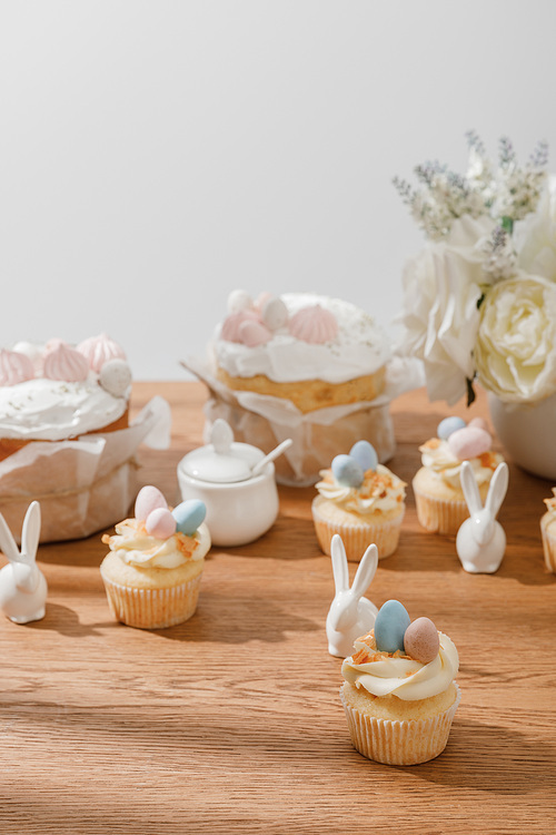 selective focus of cupcakes with decorative rabbits, sugar bowl, . cakes and vase with bouquet isolated on grey