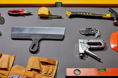 high angle view of spirit levels, pliers, measuring tape, hammer, putty knife, angle keys, stapler and tool belt on grey background