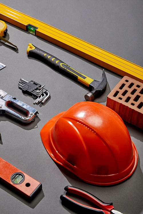 high angle view of orange helmet, hammer, spirit levels, brick, angle keys, pliers, stapler and measuring tape on grey background