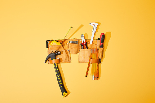 top view of tool belt with hammer, pliers, measuring tape, calipers, screwdriver and folding ruler on yellow background