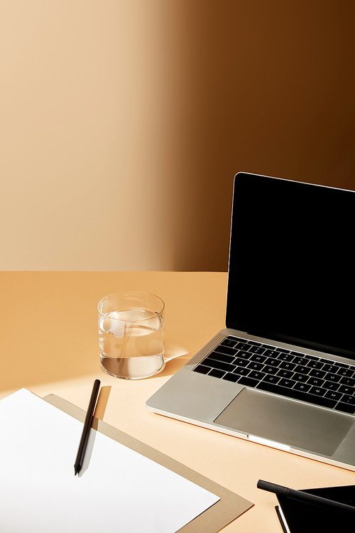 laptop with blank screen near glass of water, pencil and paper on beige surface