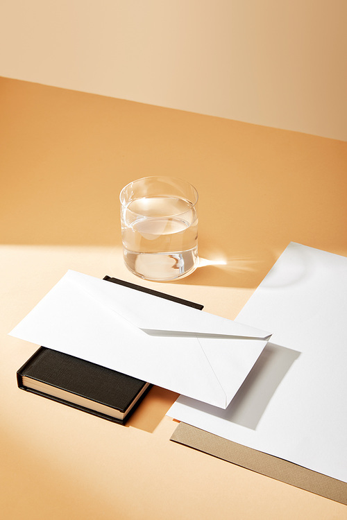 high angle view of envelope on black notebook, sheets of paper and glass of water on beige background