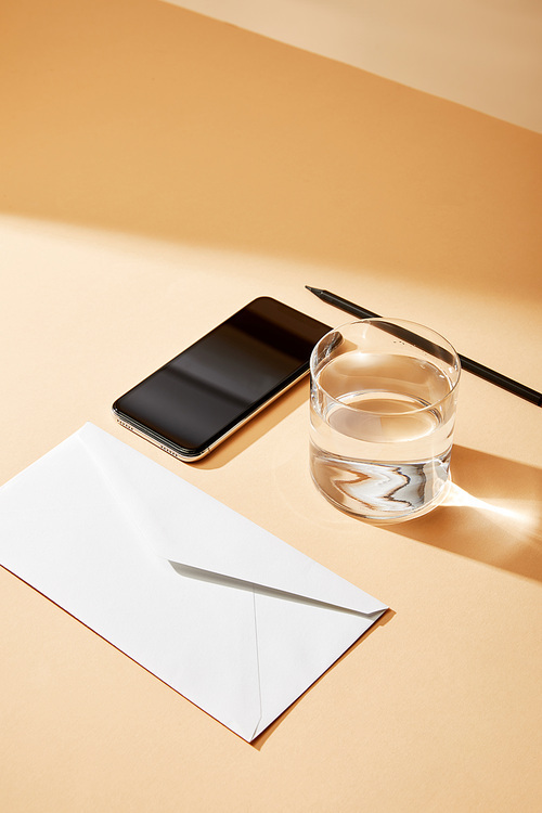 high angle view of smartphone near envelope, glass of water and pencil on beige background