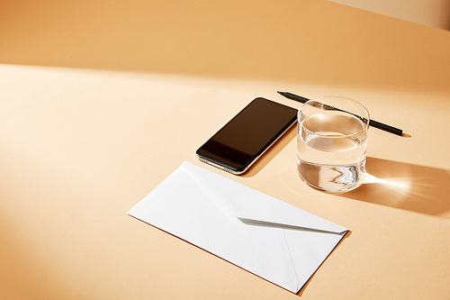 high angle view of smartphone, envelope, glass of water and pencil on beige background