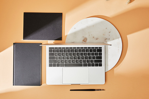 top view of laptop, marble board, black notebooks and paintbrush on beige background