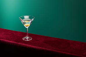 high angle view of cocktail glass with vermouth and whole olive on toothpick on velour surface on green background