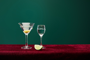 shot glass and cocktail glass with vermouth, lime slice and whole olive on toothpick on velour surface isolated on green