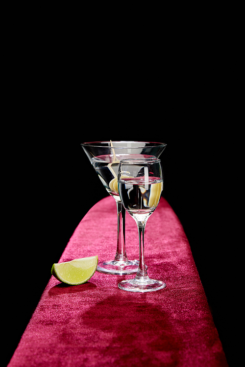 shot glass and cocktail glass with vermouth, lime slice and whole olive on toothpick on pink velour surface isolated on black