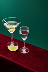 high angle view of shot glass and cocktail glass with vermouth, lime slice and whole olive on toothpick on red velour surface isolated on green