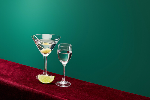 High angle view of shot glass and cocktail glass with vermouth, lime slice and whole olive on toothpick on red velour surface isolated on black